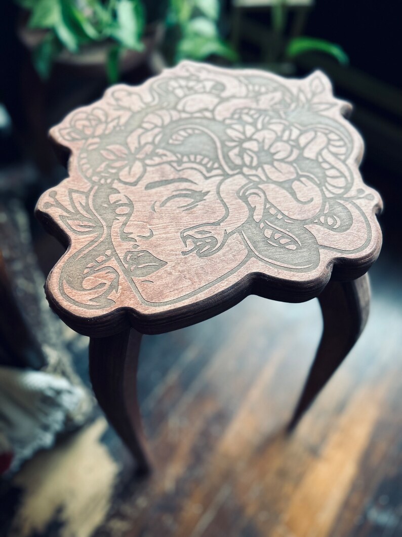 May include: A small, wooden side table with a Medusa design carved into the tabletop. The table has three curved legs and a dark brown finish. The Medusa design features a woman's face with snakes and floral elements.