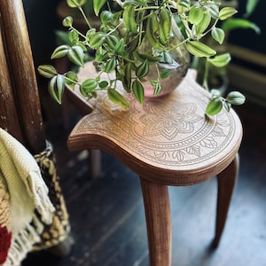 May include: A wooden side table with an intricate carved design on the top. The table has three legs and is painted a dark brown color. A green plant in a clear glass vase sits on the table.