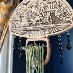 May include: A wooden mushroom-shaped wall hanging with a carved Halloween scene featuring a ghost, skull, cat, and pumpkin. Black crystal beads hang below, and a green plant is in the center. The design is etched into the wood.