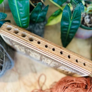 May include: A wooden yarn swift with multiple holes for holding yarn. The swift is made of light-colored wood with a detailed design. The background includes green plants and balls of yarn in shades of gray and orange.
