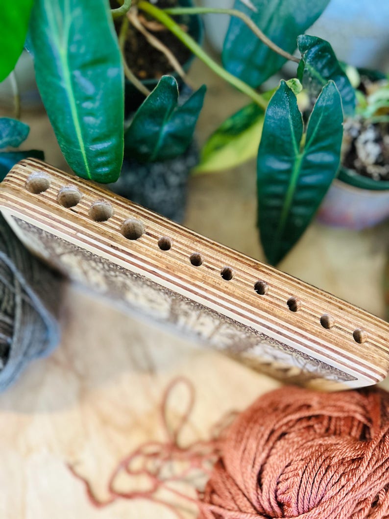 May include: A wooden yarn swift with multiple holes for holding yarn. The swift is made of light-colored wood with a detailed design. The background includes green plants and balls of yarn in shades of gray and orange.