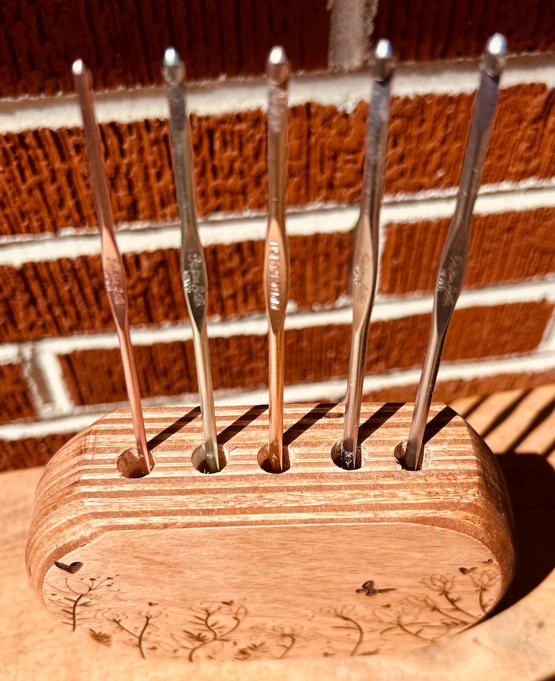 May include: A wooden stand with six slots for holding crochet hooks. The stand is made of light brown wood and has a floral design burned into the front. The crochet hooks are silver and are arranged in the slots.