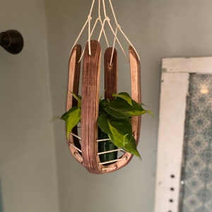 May include: A wooden hanging planter with a green plant inside. The planter is made of thin strips of wood and has a natural wood finish. The plant is a pothos, with green leaves and a trailing vine.