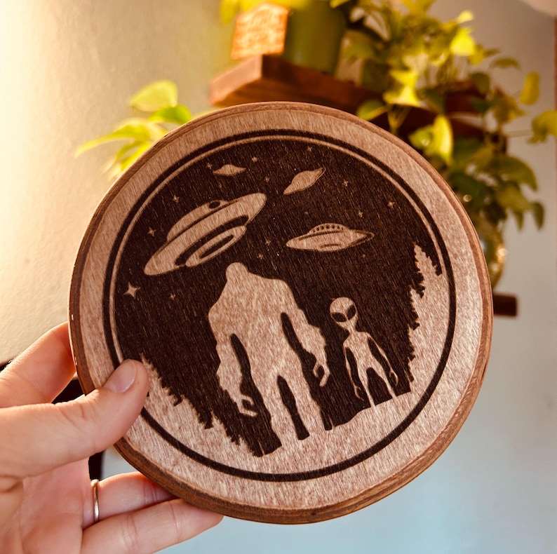 May include: A wooden round coaster with a laser-etched design of Bigfoot and an alien standing in a forest with three flying saucers in the night sky.
