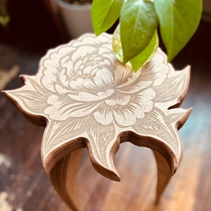 May include: A wooden side table with a floral design carved into the top. The table has three legs and is made of light brown wood.
