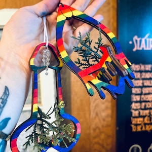 Puede incluir: Dos adornos de madera con forma de pene y de lágrima, ambos pintados con rayas de arcoíris. Los adornos están decorados con pequeñas ramitas de vegetación y una cadena de cuentas transparentes. El fondo está borroso.