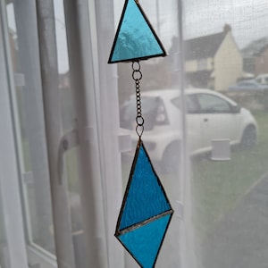 Può includere: Un acchiappasole in vetro colorato dal design geometrico. La parte superiore è un triangolo, mentre quella inferiore ha la forma di un diamante. Entrambi sono riempiti di vetro blu chiaro traslucido e collegati da una catena.