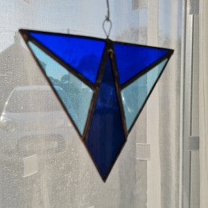 May include: A stained glass suncatcher in a geometric triangle shape. The suncatcher features shades of blue and light blue glass, framed by a dark metal border. It hangs from a metal ring and hook.