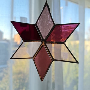 May include: A stained glass star ornament with a six-pointed design. The star features diamond-shaped panels in shades of pink, white, and burgundy, framed by a dark metal border. A white ribbon is attached for hanging.