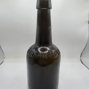 Puede incluir: Una botella de vidrio ámbar oscuro con cuello largo y base redondeada. La botella tiene el texto en relieve "Johann Hoff" en el frente. La botella está vacía y parece ser antigua.