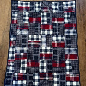 May include: A patchwork quilt with a mix of navy blue, red, and white plaid patterns. The quilt is constructed from sewn squares and has a navy blue border. It's likely made from cotton or a similar material. The quilt's dimensions are approximately 120 cm x 150 cm.