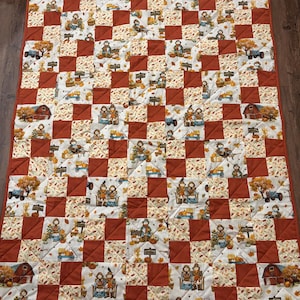 Può includere: Una trapunta fatta a mano con un design patchwork con quadrati alternati di tessuto color ruggine e tessuto color crema stampato con illustrazioni a tema autunnale. La trapunta presenta immagini di zucche, fienili e figure.