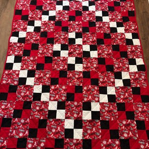 May include: A handmade quilt featuring a repeating pattern of red, black, and white squares. Some squares display the letter "A" and the word "ALABAMA" in white on a red background. The quilt is likely made of cotton and has a quilted design.
