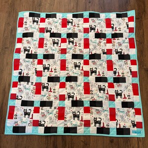 May include: A quilt with a checkered pattern featuring black, white, red, and blue squares. Each square has a different design of cats in various poses. The quilt is edged with a light blue fabric.
