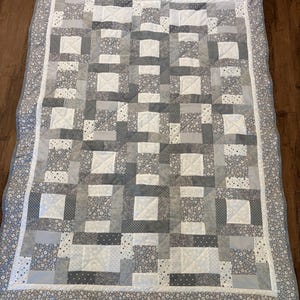 Puede incluir: Un edredón hecho a mano en tonos grises y blancos, con un diseño de patchwork. El edredón tiene un patrón repetitivo de cuadrados y rectángulos, con acentos de tela floral y de lunares. El borde es un estampado floral gris. Un textil para el hogar acogedor y decorativo.