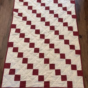 May include: A handmade quilt with a checkerboard design of alternating white squares with tiny red polka dots and solid burgundy squares. The quilt has a burgundy border and is likely made of cotton.