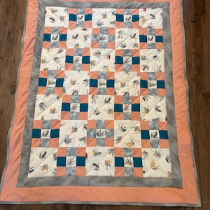 May include: A handmade quilt with a patchwork design. The quilt features a cream-coloured background with a repeating pattern of teal, peach, and grey squares. The squares are bordered by a grey and peach border.