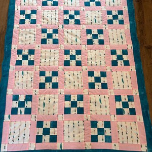 Può includere: Una trapunta fatta a mano con bordo e retro turchese. La trapunta presenta un motivo di quadrati alternati in turchese e bianco con bordi rosa. I quadrati bianchi hanno un motivo di frecce e piume.