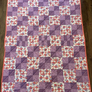 May include: A handmade quilt featuring a patchwork design with squares of varying shades of purple and white. The white squares are patterned with red and orange owls. The quilt is bordered with a coral pink binding.