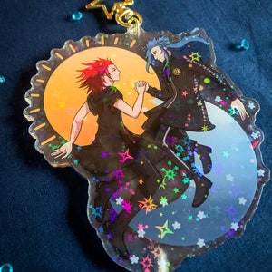 May include: A holographic charm featuring two figures, one with red hair and a black dress, the other with blue hair and a black suit, holding hands against a background of stars and a crescent moon.