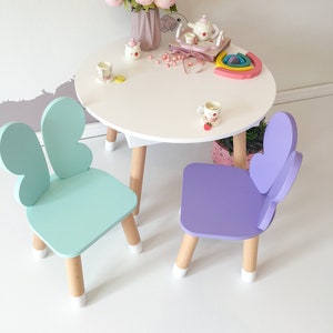 Könnte beinhalten: Ein weißer Tisch mit einem Satz Schmetterlingsstühlen in Hellblau und Lila. Der Tisch hat eine weiße Tischplatte und Holzbeine. Die Stühle haben Schmetterlingsflügel an der Rückenlehne und Holzbeine mit weißen Spitzen.