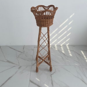 May include: A wicker plant stand with a decorative, scalloped top basket. The stand is made of natural woven wicker and has a tall, slender design.