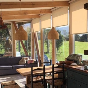 Puede incluir: Un comedor con una mesa y sillas de madera, un sof&aacute; y una cocina con armarios azules. La habitaci&oacute;n tiene grandes ventanales con persianas beige y l&aacute;mparas colgantes de mimbre.