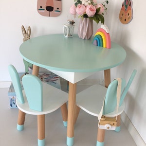 May include: A light blue children's table and two chairs with white tops and wooden legs. The table has a light blue top and the chairs have light blue backs and seats. The table and chairs have light blue accents on the legs. There is a rainbow toy and a lantern on the table.