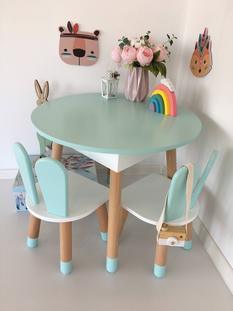 May include: A light blue children's table and two chairs with white tops and wooden legs. The table has a light blue top and the chairs have light blue backs and seats. The table and chairs have light blue accents on the legs. There is a rainbow toy and a lantern on the table.