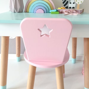 Op de afbeelding: Een roze kinderstoel met een stervormige uitsparing in de rugleuning. De stoel heeft vier houten poten met lichtblauwe geverfde uiteinden. De stoel staat op een wit oppervlak.