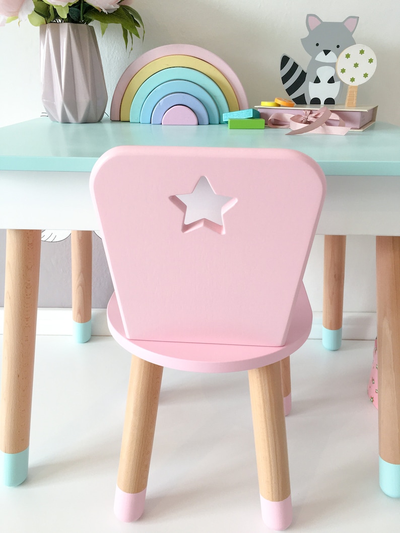 Op de afbeelding: Een roze kinderstoel met een stervormige uitsparing in de rugleuning. De stoel heeft vier houten poten met lichtblauwe geverfde uiteinden. De stoel staat op een wit oppervlak.
