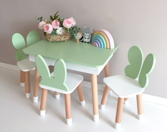 May include: A green children's table and four chairs with bunny ears. The chairs are white with natural wood legs. The table has a green top and natural wood legs.