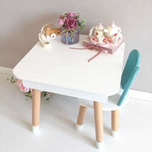 Op de afbeelding: Witte houten kindertafel en stoel set met een turquoise stoel. De tafel heeft een wit blad en houten poten met witte accenten. Een roze bloemenarrangement staat op de tafel met een houten speelgoedcamera en een theeset.