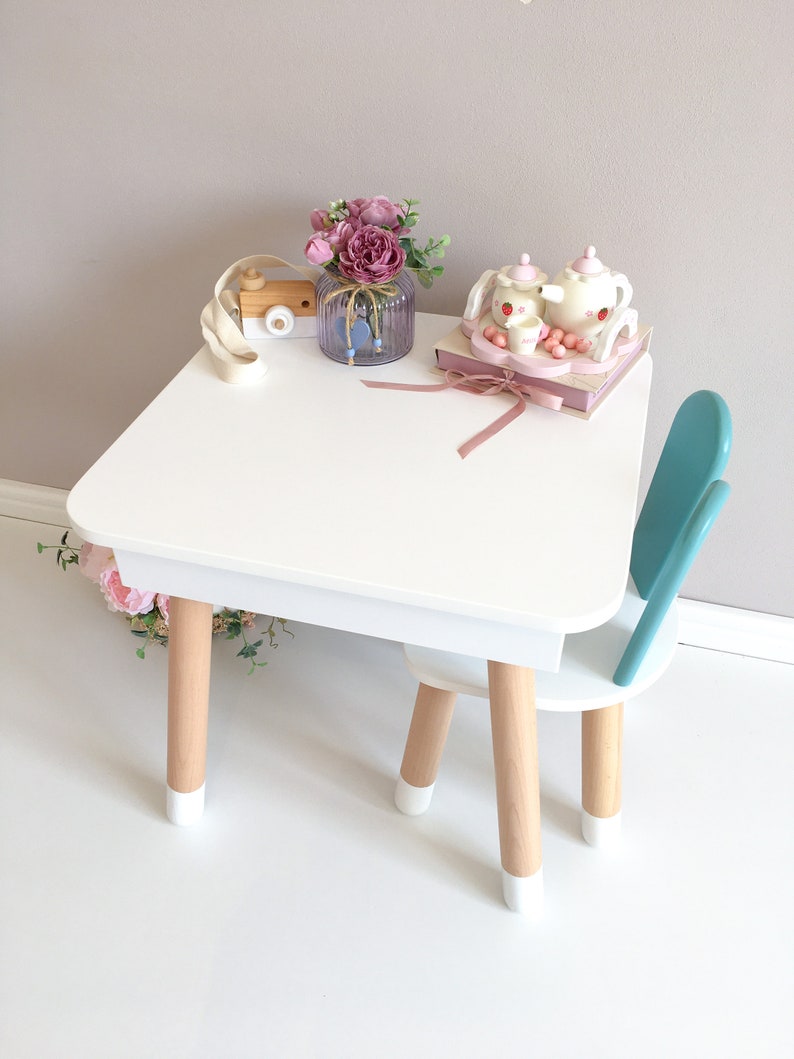 Op de afbeelding: Witte houten kindertafel en stoel set met een turquoise stoel. De tafel heeft een wit blad en houten poten met witte accenten. Een roze bloemenarrangement staat op de tafel met een houten speelgoedcamera en een theeset.