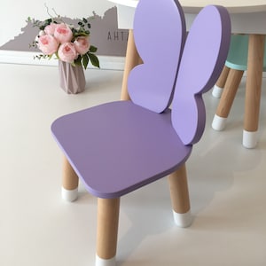 May include: A purple butterfly-shaped children's chair with wooden legs and white painted feet. The chair is on a white surface.