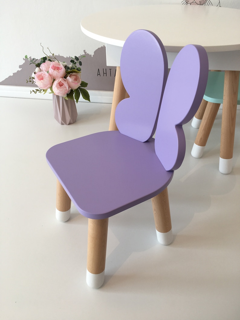 May include: A purple butterfly-shaped children's chair with wooden legs and white painted feet. The chair is on a white surface.