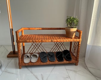 May include: A two-tiered wicker shelf with a woven design. The shelf is brown and has a small potted plant on the top shelf. There are four pairs of slippers on the bottom shelf, two pairs are gray and two pairs are black.