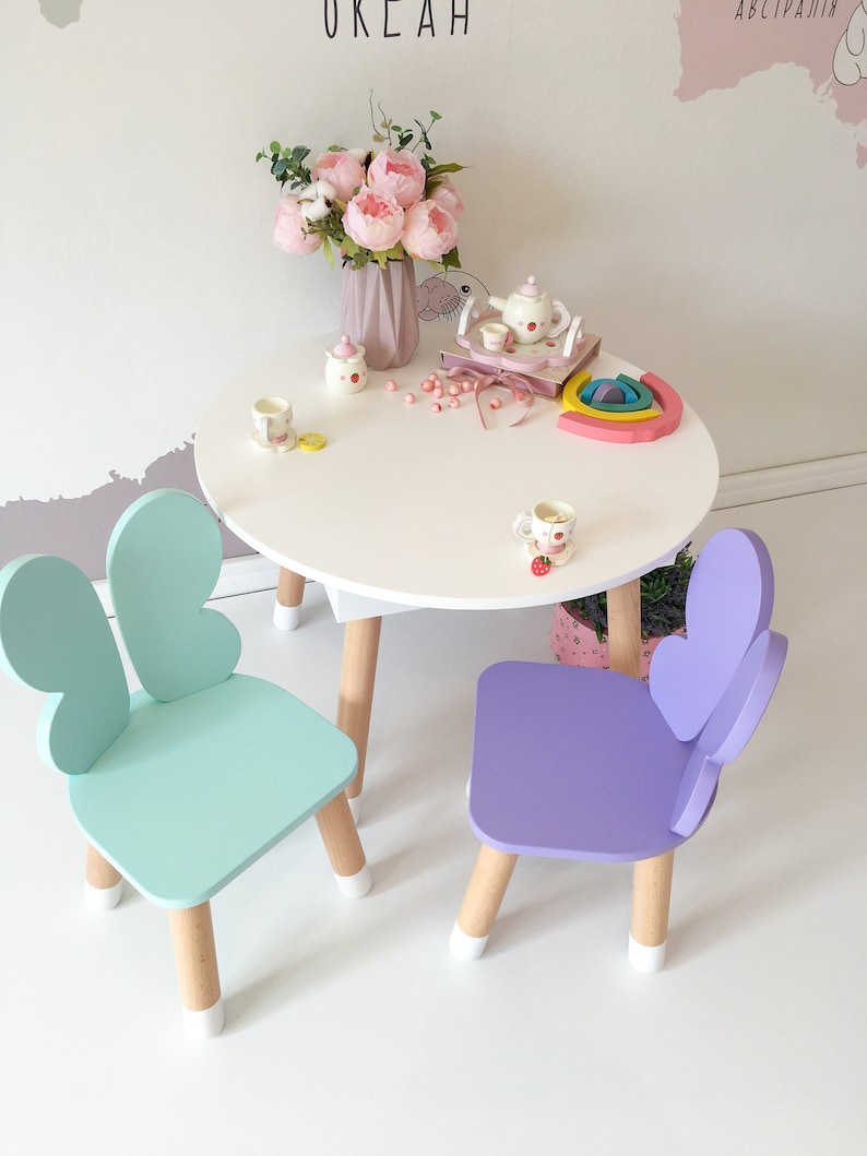 May include: A white table with a light blue and purple butterfly chair set. The table has a white top and wooden legs. The chairs have butterfly wings on the back and wooden legs with white tips.
