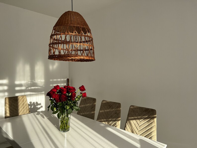 May include: A white dining table with a wicker pendant light hanging above it.  There are four chairs around the table, and a vase of red roses in the center.
