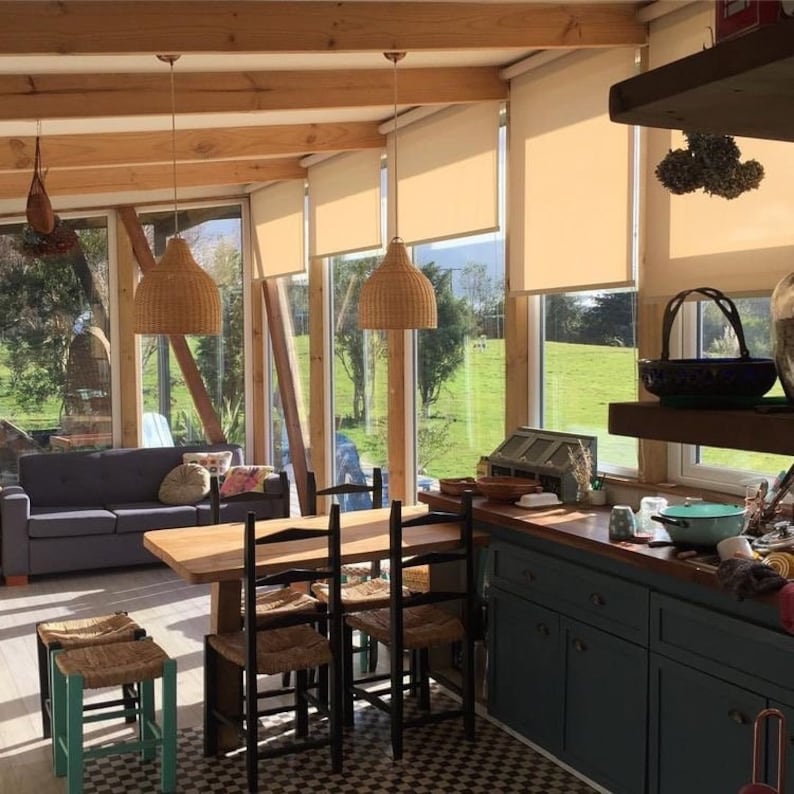 Puede incluir: Un comedor con una mesa y sillas de madera, un sof&aacute; y una cocina con armarios azules. La habitaci&oacute;n tiene grandes ventanales con persianas beige y l&aacute;mparas colgantes de mimbre.