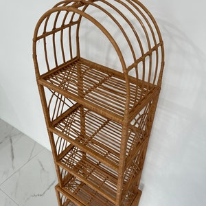 May include: A tall, natural woven rattan shelf unit with four shelves and an arched top. The shelf unit is made of light brown rattan and has a natural finish.