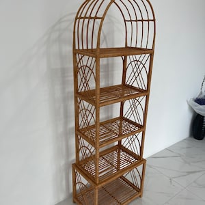 May include: A tall, natural woven rattan shelf unit with an arched top and four shelves. The shelf unit is made of light brown rattan and has a natural finish.