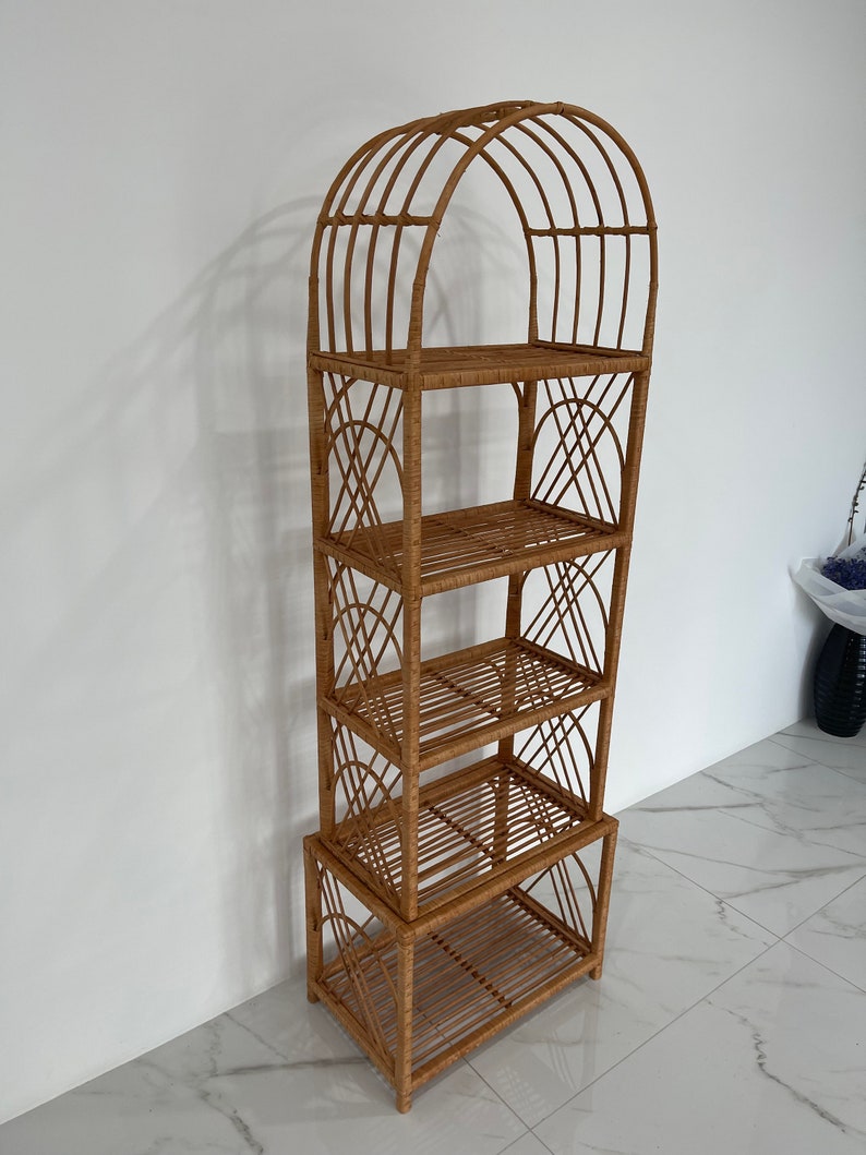 May include: A tall, natural woven rattan shelf unit with an arched top and four shelves. The shelf unit is made of light brown rattan and has a natural finish.