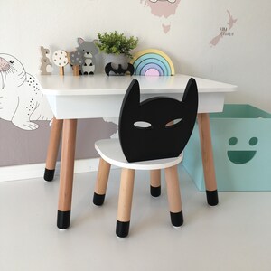 Op de afbeelding: Een witte houten tafel en stoel set met zwarte accenten. De stoel heeft een zwarte Batman-vormige rugleuning. De tafel heeft een regenboogkleurige houten boog en een verscheidenheid aan houten dierenfiguren erop.
