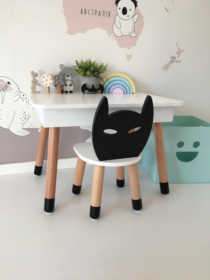 Op de afbeelding: Een witte houten tafel en stoel set met zwarte accenten. De stoel heeft een zwarte Batman-vormige rugleuning. De tafel heeft een regenboogkleurige houten boog en een verscheidenheid aan houten dierenfiguren erop.