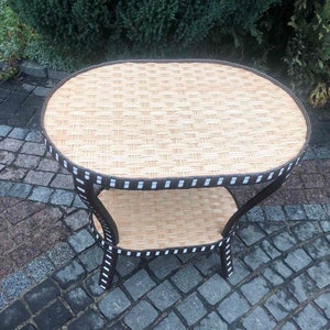 May include: A brown wicker and metal oval-shaped side table with two tiers. The table has a black and white trim around the edges.