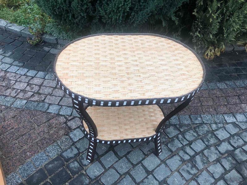 May include: A brown wicker and metal oval-shaped side table with two tiers. The table has a black and white trim around the edges.