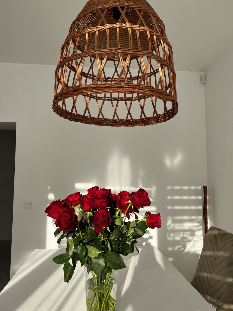 May include: A bouquet of deep red roses in a clear glass vase on a white table. The roses are in focus, while the background is blurred. A wicker lampshade hangs from the ceiling above the table.