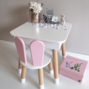 Op de afbeelding: Een witte tafel en roze stoel set met houten poten. De tafel heeft een wit blad en een roze lade. De stoel heeft een roze rugleuning en zitting. Er staan decoratieve items op de tafel, waaronder een vaas met witte bloemen, een wasbeer figuur, een konijn figuur en een roze doos met het woord "Cactus" erop.