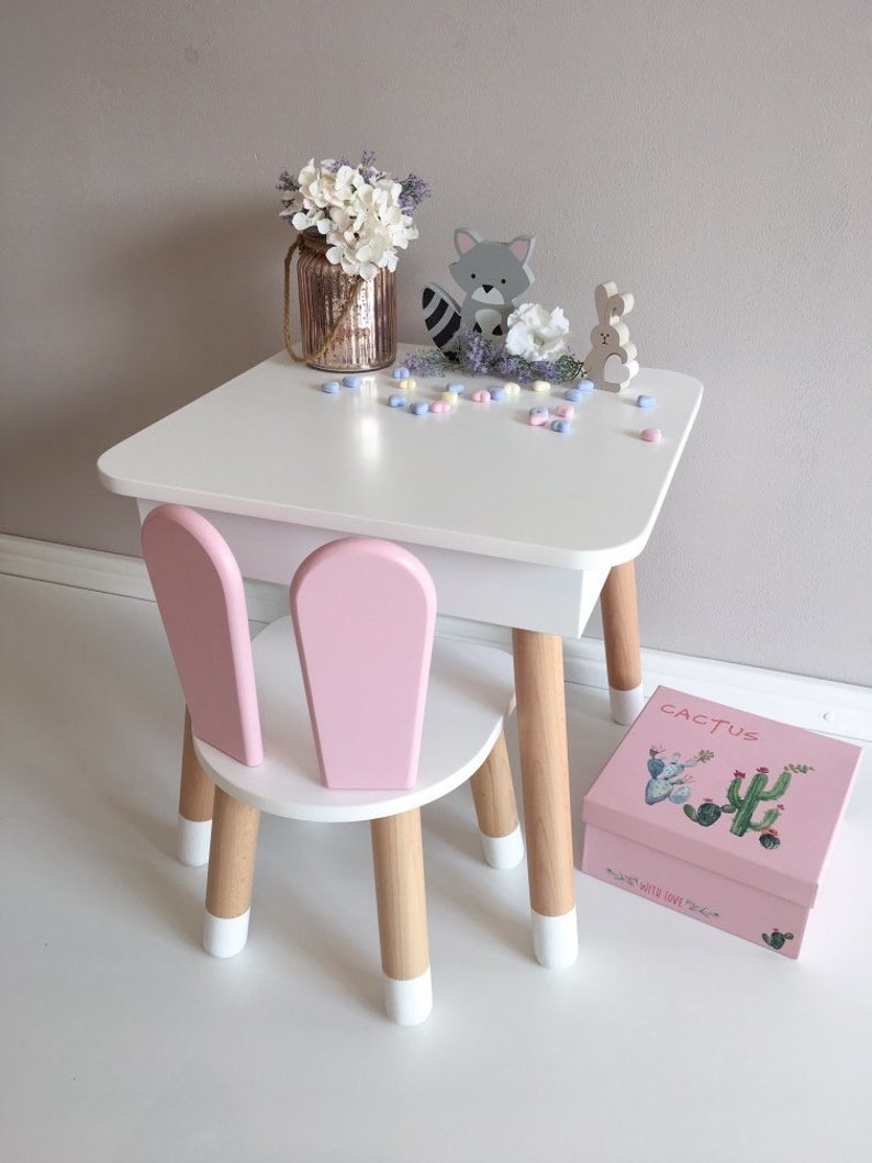Op de afbeelding: Een witte tafel en roze stoel set met houten poten. De tafel heeft een wit blad en een roze lade. De stoel heeft een roze rugleuning en zitting. Er staan decoratieve items op de tafel, waaronder een vaas met witte bloemen, een wasbeer figuur, een konijn figuur en een roze doos met het woord "Cactus" erop.